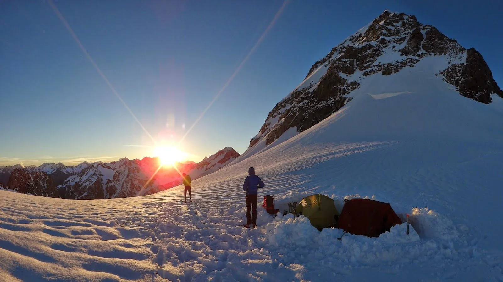 Two figures at a snow camp at sunrise with a tent, a peak behind
