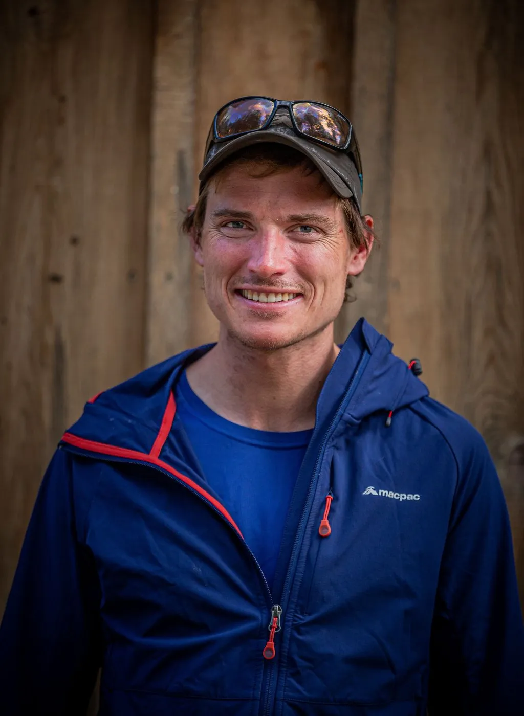 Portrait of Alastair McDowell in a blue jacket and cap against a timber wall