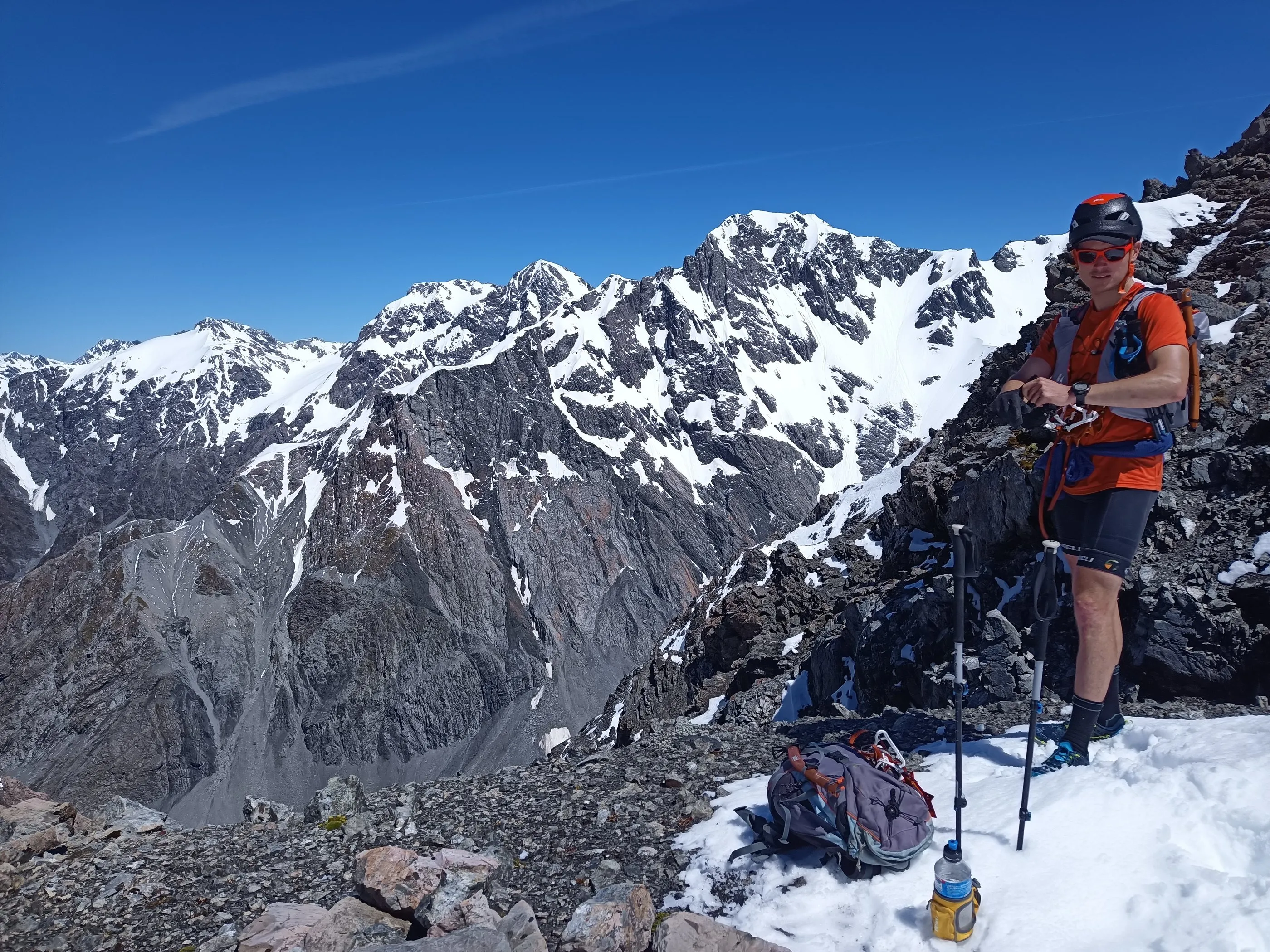 Alastair McDowell standing on a rocky ridge with poles, the snow-covered Main Divide behind him