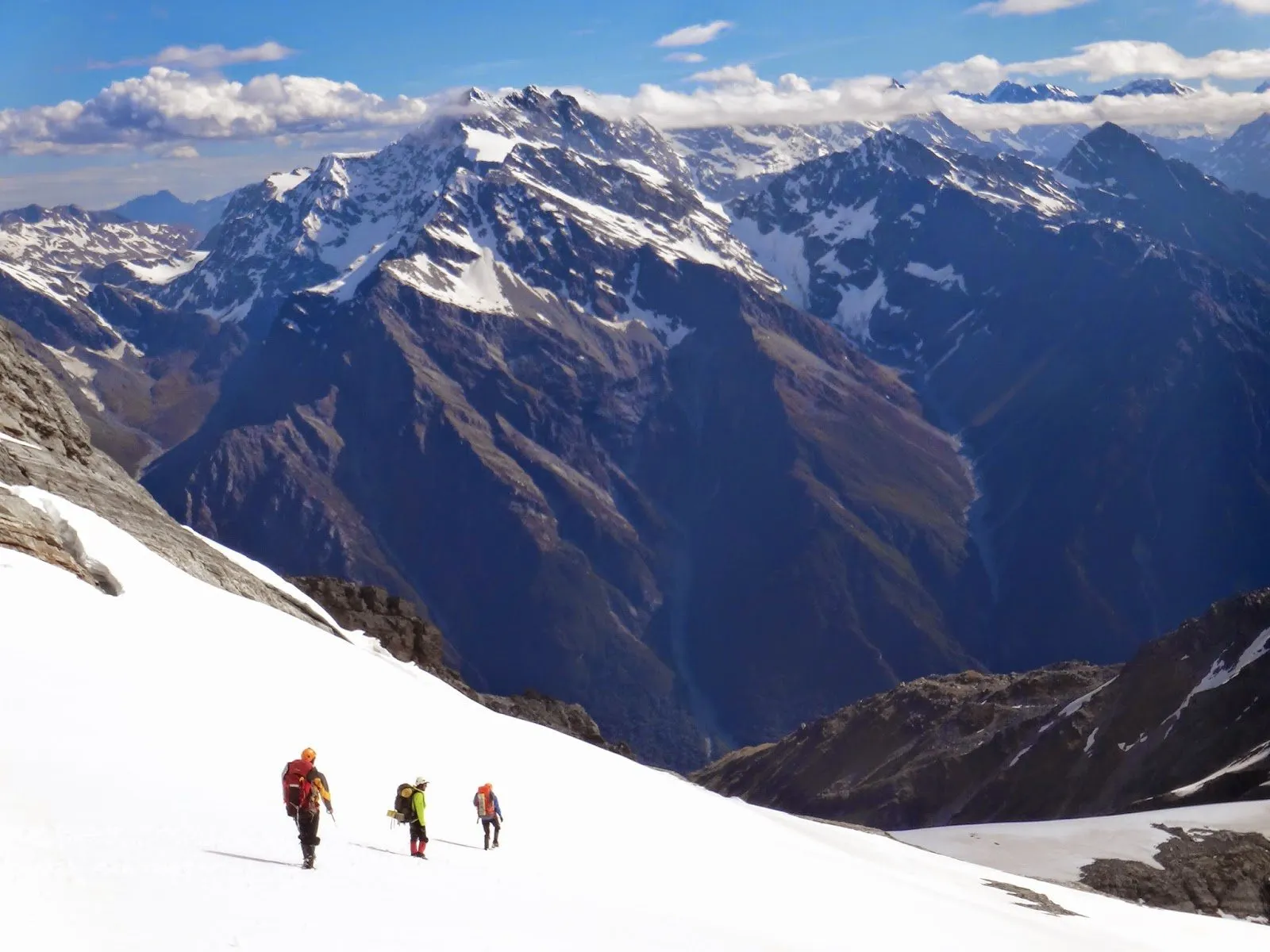 Three climbers crossing a snow slope with a range of peaks beyond